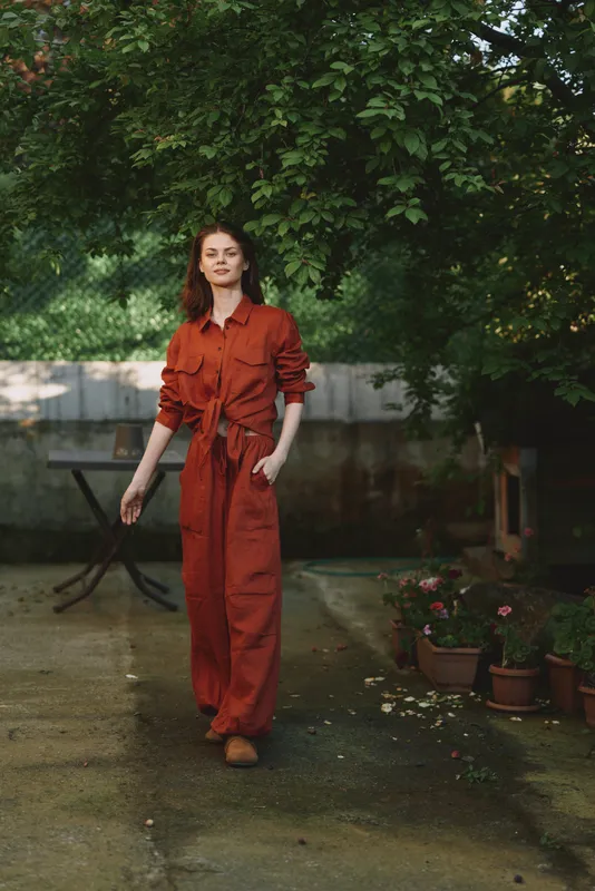 Woman in orange jumpsuit stands in a garden setting, enjoying a relaxed lifestyle moment, casual pose beside potted plants, natural light highlights contemporary fashion in an outdoor scene