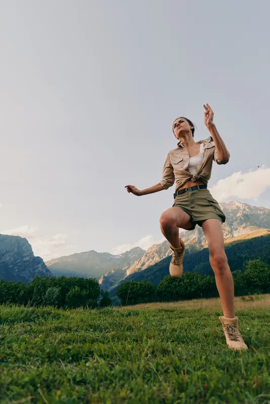 Woman jumping in a meadow with mountains in the background, casual outdoor attire, bright sky, and grass underfoot, capturing a vibrant lifestyle and energetic outdoor moment.