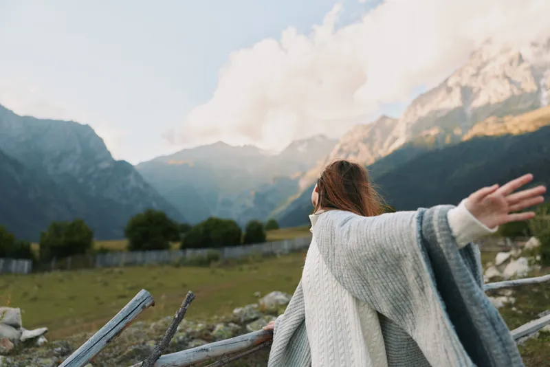 A woman stands outdoors with arms outstretched, facing mountains and a sunlit landscape, conveying freedom, joy, and a relaxed lifestyle in nature.