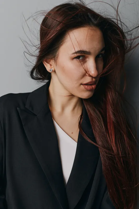 Woman with long hair and serious expression wearing black blazer and white top posing against gray background. Portrait of confident female with natural makeup and loose hairstyle.