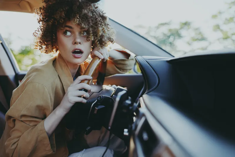 Young woman sits in a car interior with a surprised expression, casual fashion, natural light, retro film color tones, candid lifestyle moment, isolated on empty road in frame style.