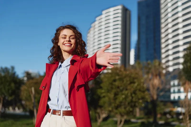 Young woman in a red coat raises her arms with a joyful smile in a sunlit urban park, modern glass towers behind her and green trees in the foreground
