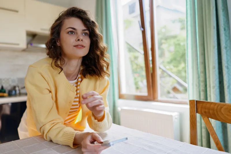 Casual lifestyle portrait of a thoughtful woman at home, seated at a dining table by a window, wearing a yellow cardigan and striped shirt, hand on a pen, calm focused mood, natural light.