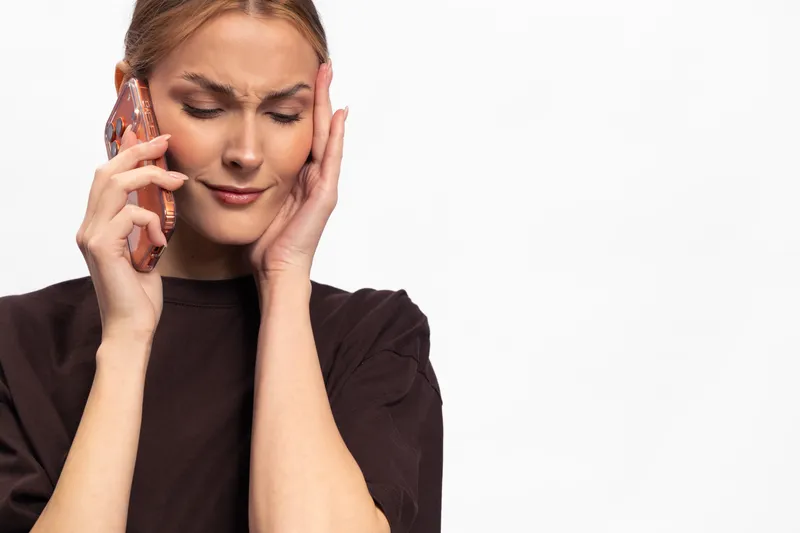 Stressed young woman in dark clothing holds phone to ear and touches her temple with concerned expression. Isolated on white background. Concept for anxiety, headache, and communication issues.