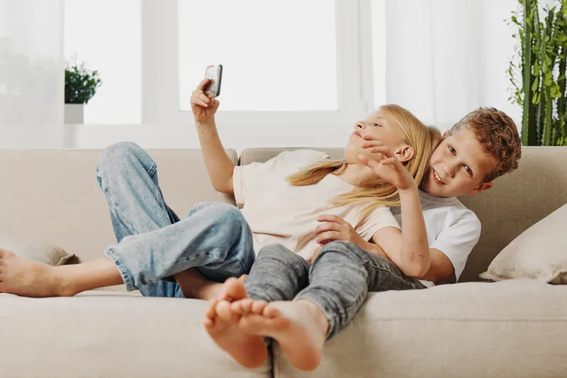 Two happy children lying on sofa taking selfie together in bright living room. Casual home environment with smiling boy and girl enjoying playful moment and modern family lifestyle.