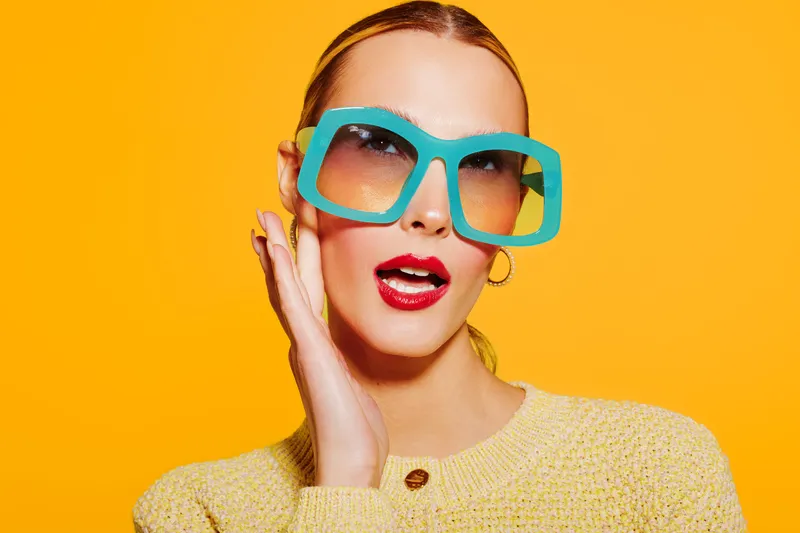 Stylish woman with large blue sunglasses and red lipstick poses against bright yellow background. Fashion concept with bold colors and confident expression in studio lighting.