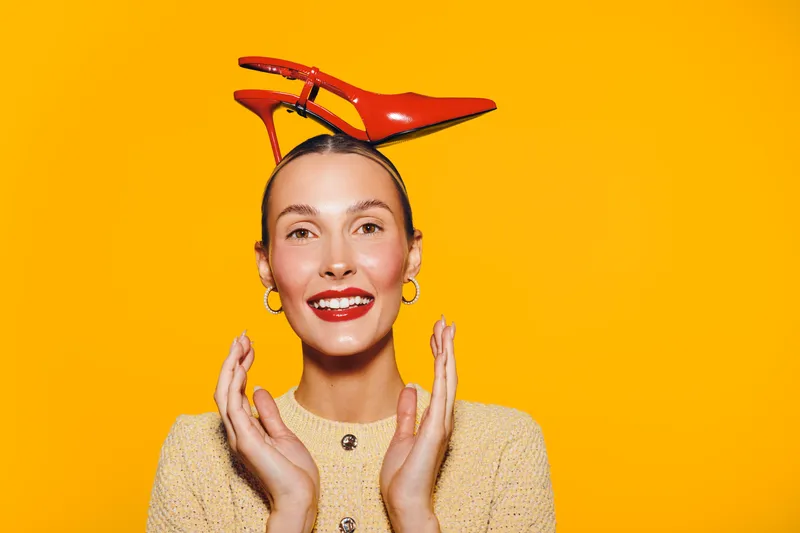 Smiling woman with red lipstick and earrings balances red high heel shoes on her head against bright yellow background. Concept of fun, fashion, and playful mood.
