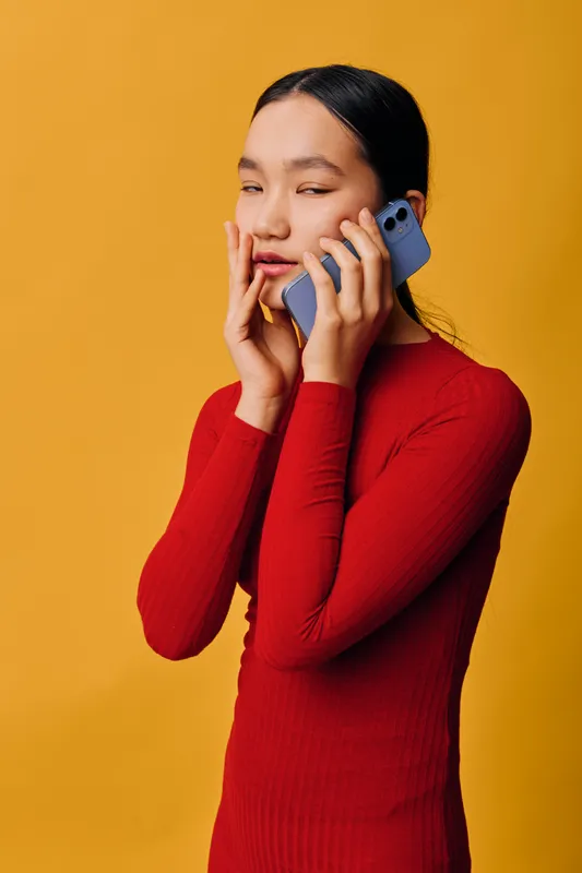 Woman in red sweater talking on smartphone with thoughtful expression, holding hand near face isolated on yellow background. Portrait of young female using mobile phone communication.