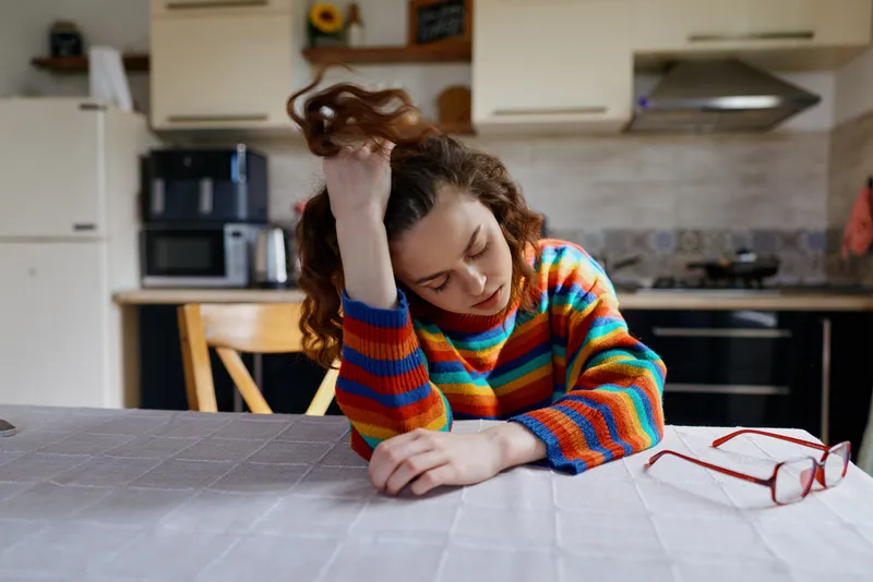 Woman with curly hair rests her head on her hand at a table in a bright kitchen, wearing a colorful striped sweater. Casual home lifestyle, relaxed mood, soft daylight, everyday moment.