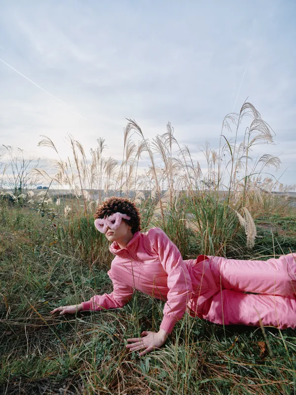 A person in a pink outfit lies on grass in an open field, relaxed pose among tall weeds, blue sky and soft light creating a casual outdoor portrait with nature