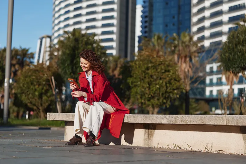 Woman with curly hair wearing a red coat sits on a low concrete bench in an urban park, absorbed in her smartphone as modern buildings rise behind trees. Soft daylight highlights casual street style