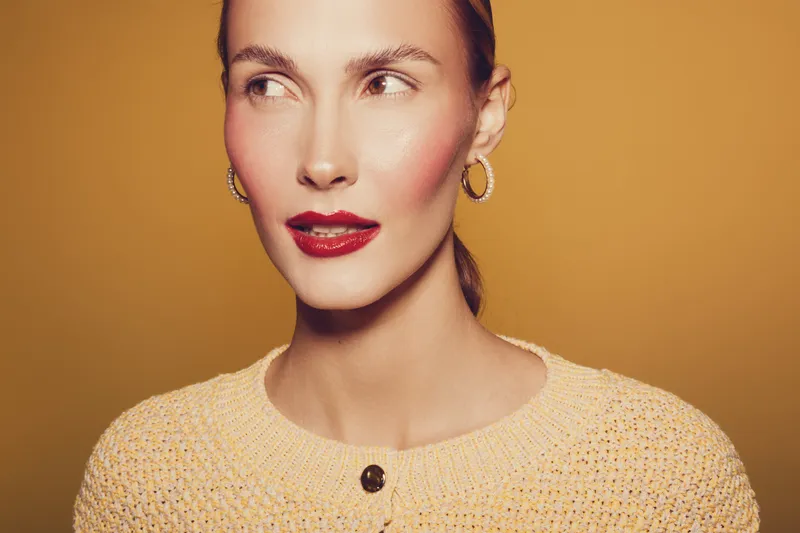 Portrait of a woman with red lipstick and hoop earrings wearing a knitted sweater on a warm brown background. Concept of beauty, fashion, and confident female model.