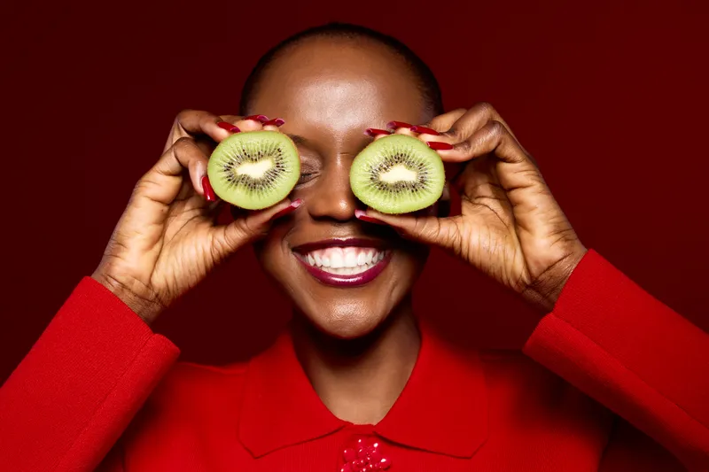 Dark skinned African woman with short hair smiles holding two kiwi halves over her eyes wearing red outfit. Concept for healthy lifestyle, fruit, wellness, and joyful natural beauty.
