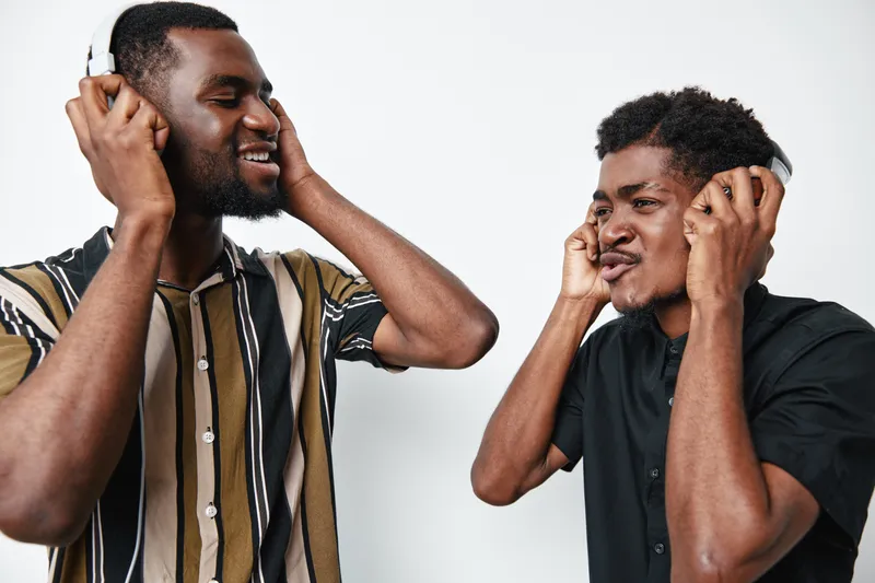 Two young black men with dark skin wearing headphones enjoying music together, smiling and interacting in casual clothing against white background in studio setting.