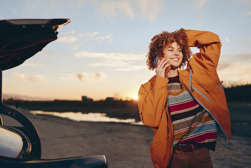 Woman lifestyle outdoors near car at sunset, curly hair in orange jacket, smiling and adjusting hood, casual travel mood, open road background, sunlit sky, warm natural light, evening