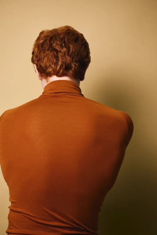 Person with red hair and brown sweater seen from behind against beige background. Back view, casual clothing and natural hair color with soft lighting and warm tones.