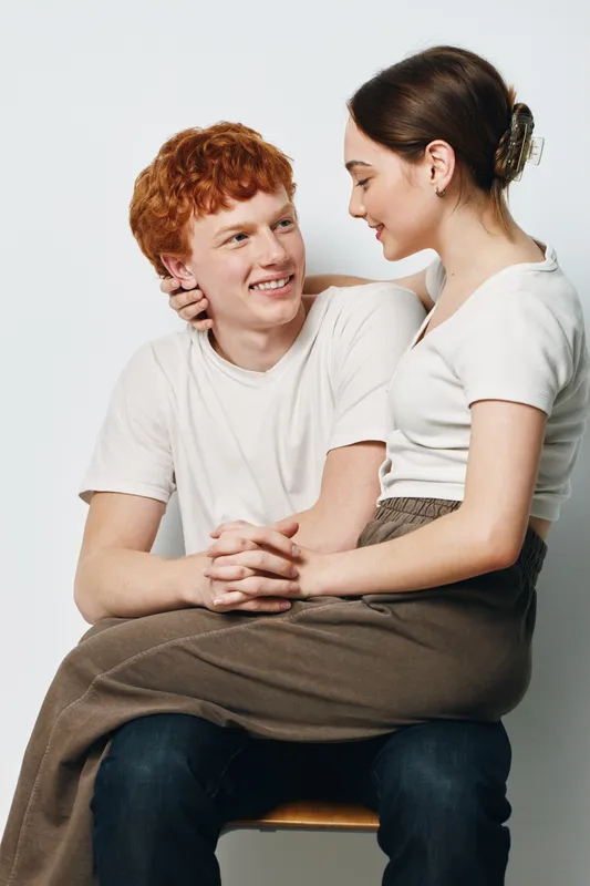 Couple sitting together with woman on man’s lap, smiling and looking at each other on white background in casual clothes. Young people enjoying moment of closeness and affection indoors.