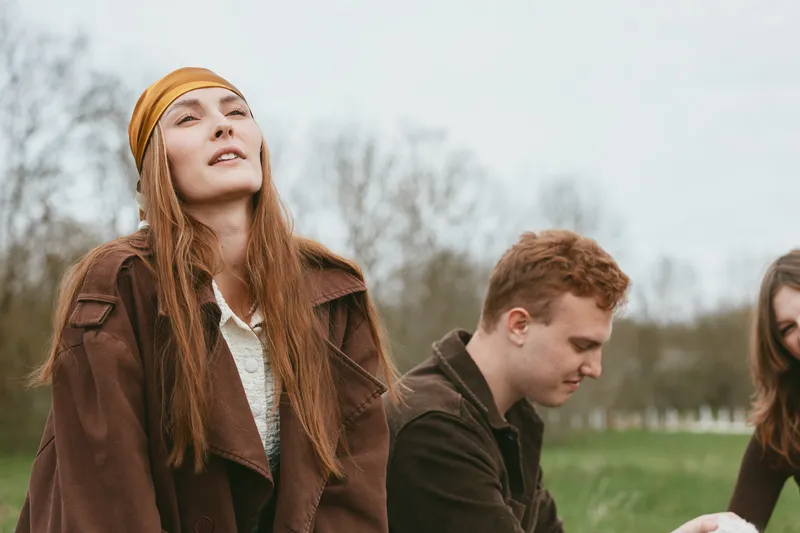 Outdoor lifestyle scene with a smiling woman in a brown coat and headband, surrounded by friends in a park. Natural light captures a warm, candid mood and relaxed interaction outdoors.
