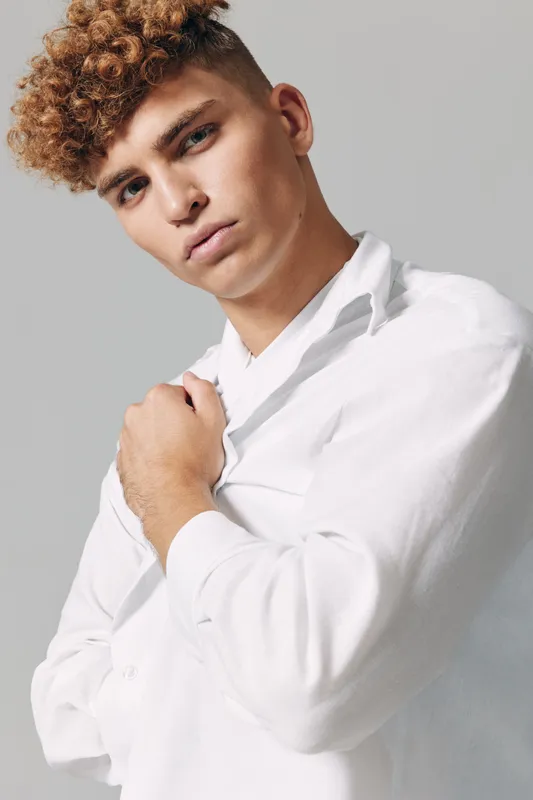 Young man with curly hair wearing white shirt posing with serious expression on gray background in studio portrait fashion style.