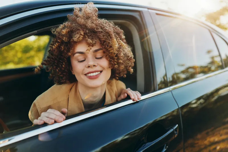 Young woman leans from a car window, smiles with curly hair, enjoying an outdoor drive in casual fashion, captured with vintage retro film color for a warm lifestyle moment.