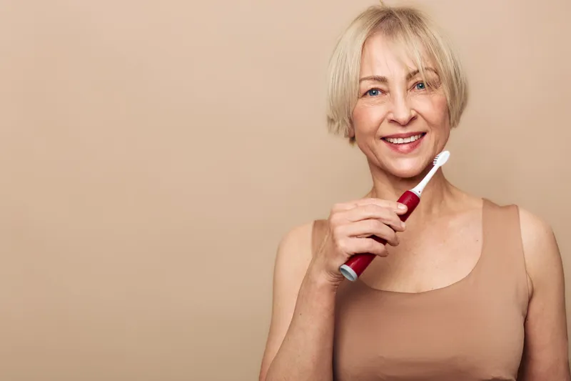 Smiling middle aged woman with short blonde hair holds electric toothbrush near face. Healthy teeth and oral hygiene concept. Neutral beige background highlights natural beauty and wellness.