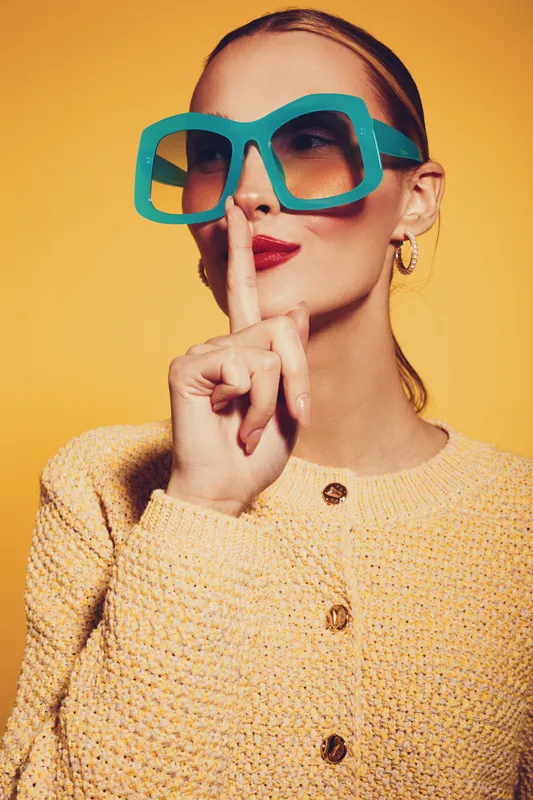 Stylish woman in oversized turquoise sunglasses and yellow knitted sweater makes shushing gesture with finger on lips against bright yellow background. Concept of secrecy and silence.