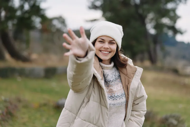 Woman in a warm winter coat reaching out with a smile in an open park, cold weather, soft natural light, candid portrait conveying friendliness and outdoor joy.