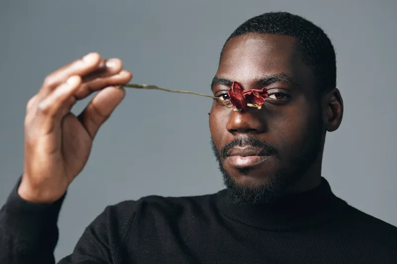 African man with dark skin holding a spoon with red flower close to his face in creative conceptual visual style. Portrait, artistic, black sweater and gray background.