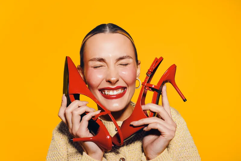 Happy woman with closed eyes holds red high heel shoes near face smiling broadly. Bright yellow background highlights joyful mood and fashion concept with stylish footwear and beauty.