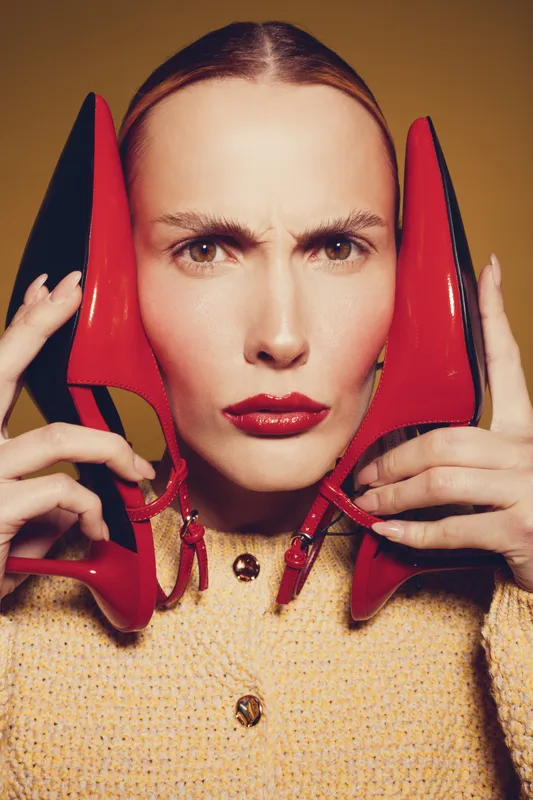 Woman with red lipstick holds red high heel shoes near face, wearing yellow sweater. Close-up portrait with serious expression on beige background. Concept of fashion and style.