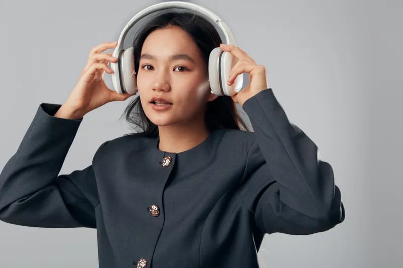 Young woman with headphones dressed in formal dark jacket listening to music or podcast with focused expression on gray background for technology and entertainment concept