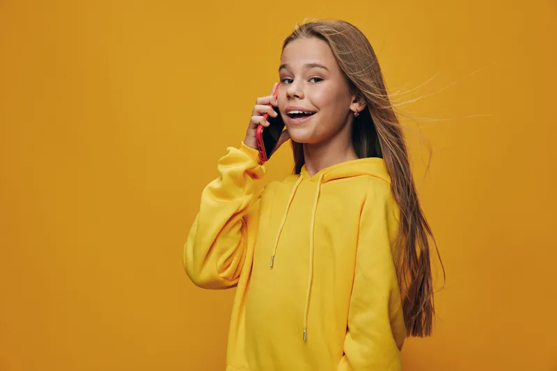 Girl with long hair wearing yellow hoodie talking on smartphone smiling happily with bright background indoor. Child expressing joy communication and youth technology portrait.