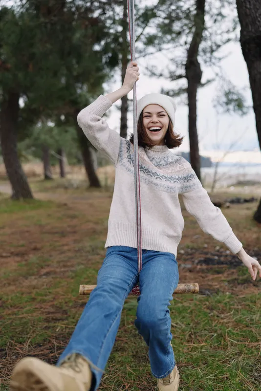 Woman on a rope swing outdoors in a forest, smiling and enjoying a playful moment in nature beneath trees and a clear sky.
