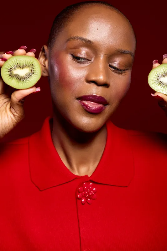 Close-up of dark skinned African woman holding two kiwi halves near her face with closed eyes. Red background and red clothing create a vibrant contrast. Concept of healthy lifestyle.