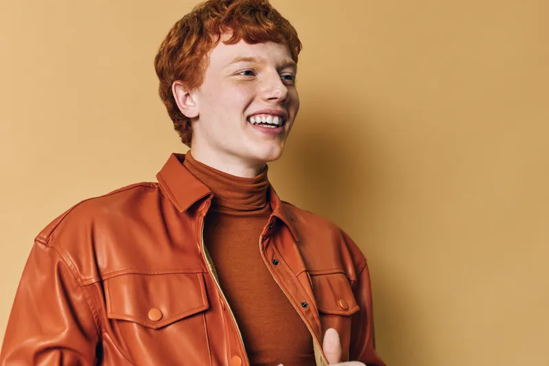 Young man with curly red hair smiling and looking away dressed in brown turtleneck and orange leather jacket standing against beige background with cheerful expression.