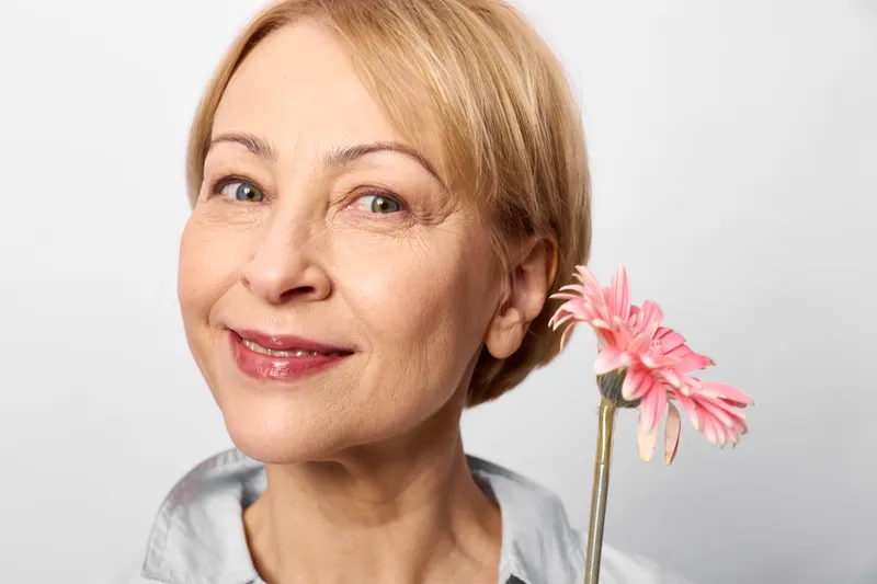 Smiling elderly woman portrait, blonde short hair, holding a pink gerbera flower, clean white background, gentle natural expression, closeup studio lighting, calm mood