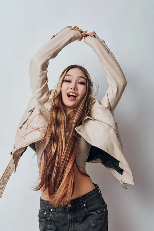 Young woman with long hair wearing casual beige jacket and black pants posing with arms raised smiling joyfully on light background. Lifestyle, fashion and happiness concept.