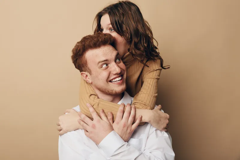 Happy young couple embraces and smiles in studio with beige background. Woman hugs man from behind showing affection and joy. Concept of love, relationship, and togetherness.