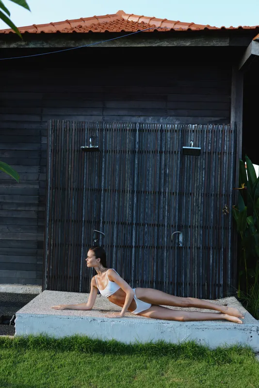 Yoga pose by a smiling woman outdoors on a mat beside a rustic wooden shed, sunlight on skin, showcasing fitness, wellness, and a calm lifestyle.
