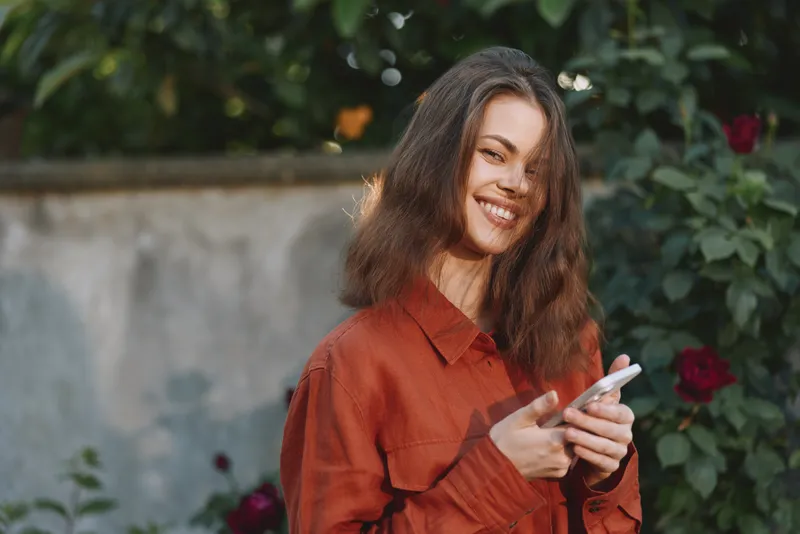 Smiling woman in a red shirt outdoors, holding a smartphone in a garden setting. A warm lifestyle portrait capturing everyday moments and casual connection!