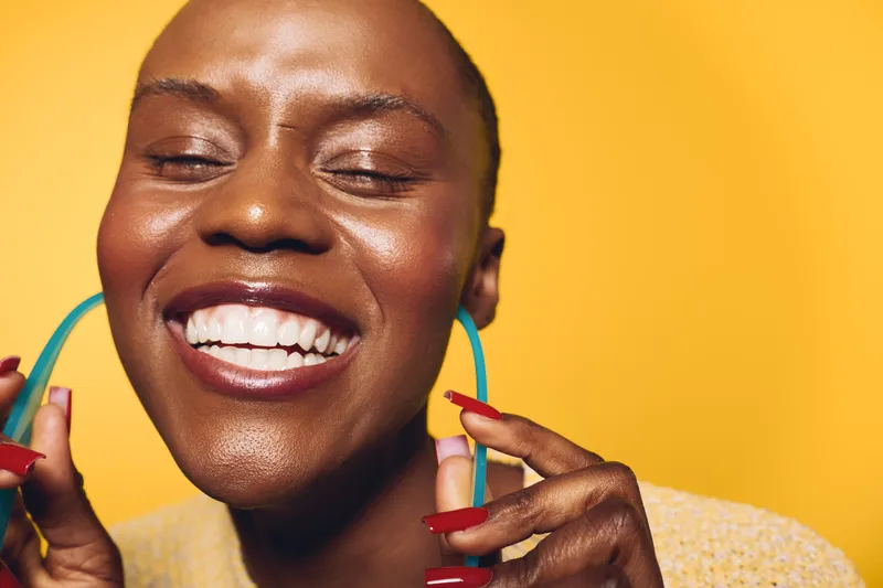 Happy African woman with dark skin smiles brightly, holding large blue earrings near her face. Close-up on yellow background. Concept for beauty, joy, and self-expression.