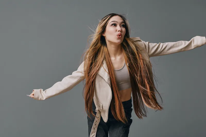 Young woman with long hair wearing casual clothing and posing enthusiastically against a gray background. Female model expressing joy and freedom in studio shot.