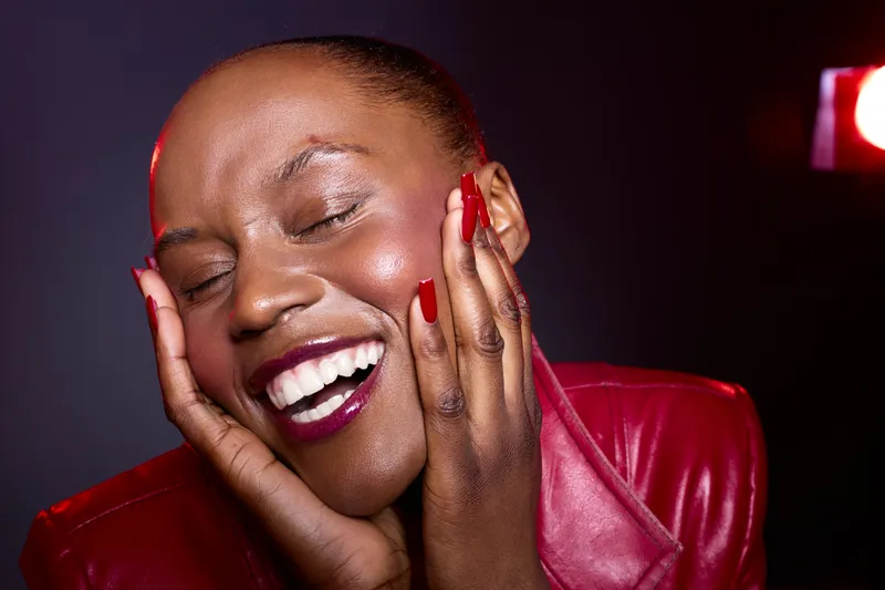 Happy African woman with dark skin smiles with closed eyes and red nails, wearing a red leather jacket. Studio portrait with dramatic lighting and joyful expression concept.