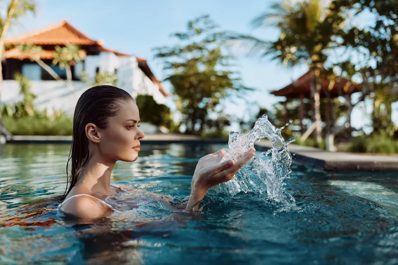 Lifestyle shot of a woman in a swimming pool at a tropical resort, creating a water splash with her hand, sunny day, calm expression, relaxing and playful mood, outdoor leisure scene.