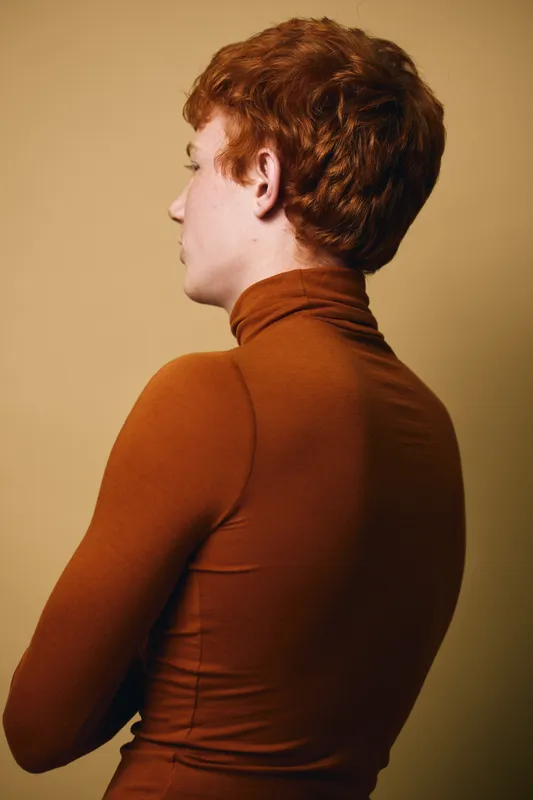 Portrait of a young man with red hair wearing a brown turtleneck sweater standing sideways against a neutral background. The image shows profile and casual style with soft lighting.