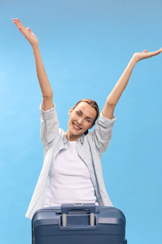 Happy young woman with raised arms stands behind suitcase on blue background. Concept for travel, vacation, freedom, and joyful journey with luggage and positive emotion.