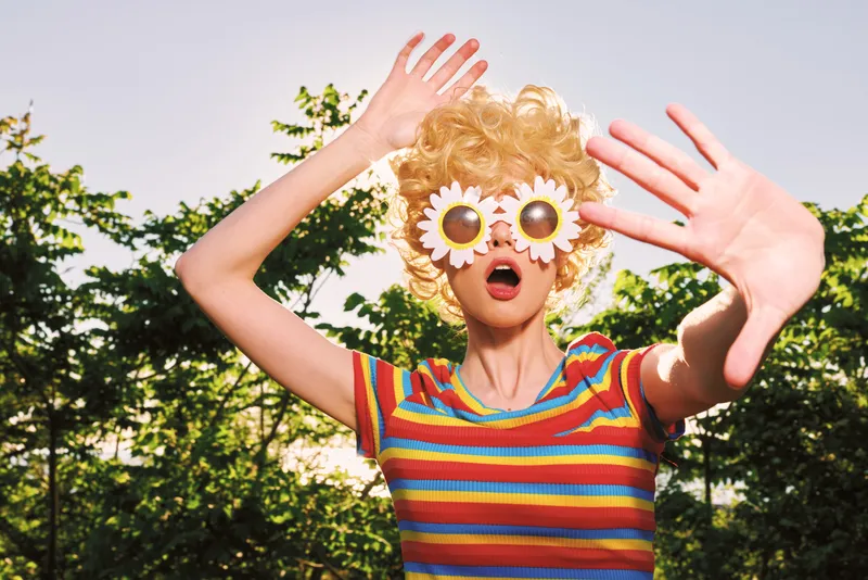 A playful outdoor scene shows a woman in a striped shirt wearing oversized daisy sunglasses, capturing a retro lifestyle moment full of color, joy, and carefree creativity.