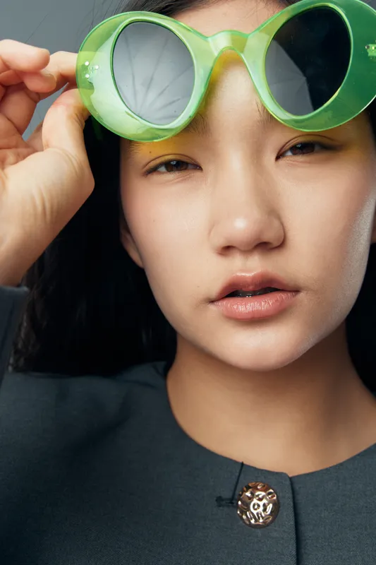 Young woman with clear skin holding green retro sunglasses near face in studio. Fashion portrait with natural makeup, black hair, and stylish clothing closeup for beauty concept.