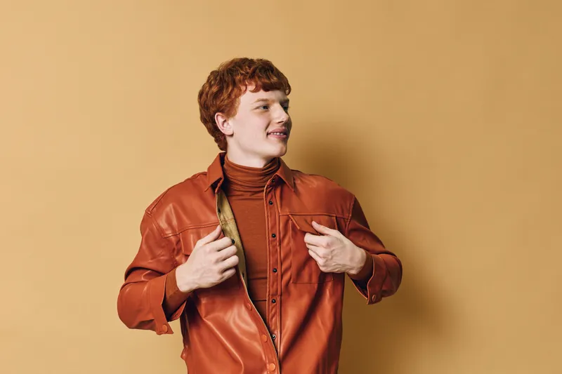 Young man with red hair wearing a brown leather jacket and turtleneck posing confidently against a beige background looking to the side with a slight smile and relaxed posture.