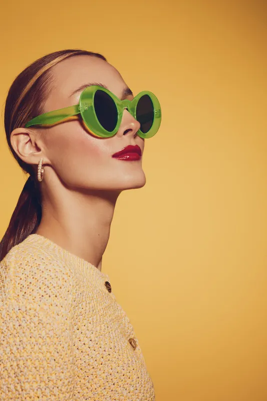Stylish woman with green round sunglasses and red lipstick poses on yellow background. Fashion portrait with retro vibe and confident expression in bright studio lighting.